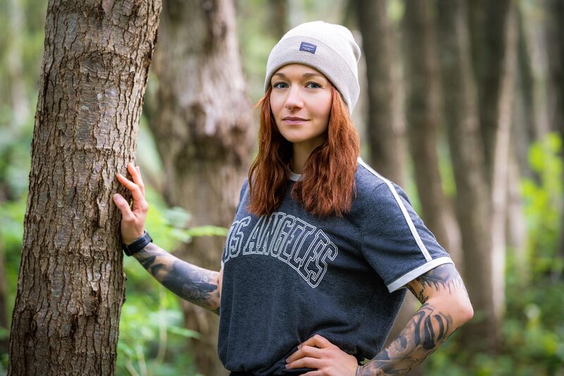 Aniko Heuert im Wald, eine Hand an einem Baumstamm, mit heller Mütze und dunklem Shirt